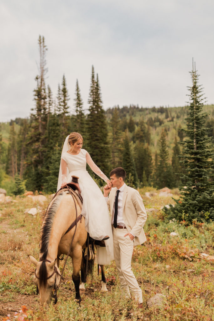 Groom plants a kiss on bride's hand as she sits on horse, taken by Utah western wedding photographer Robin Kunzler