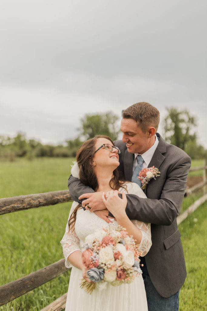 Groom hugs bride from behind as they smile at each other by wood post fence, taken by Utah western wedding photographer Robin Kunzler