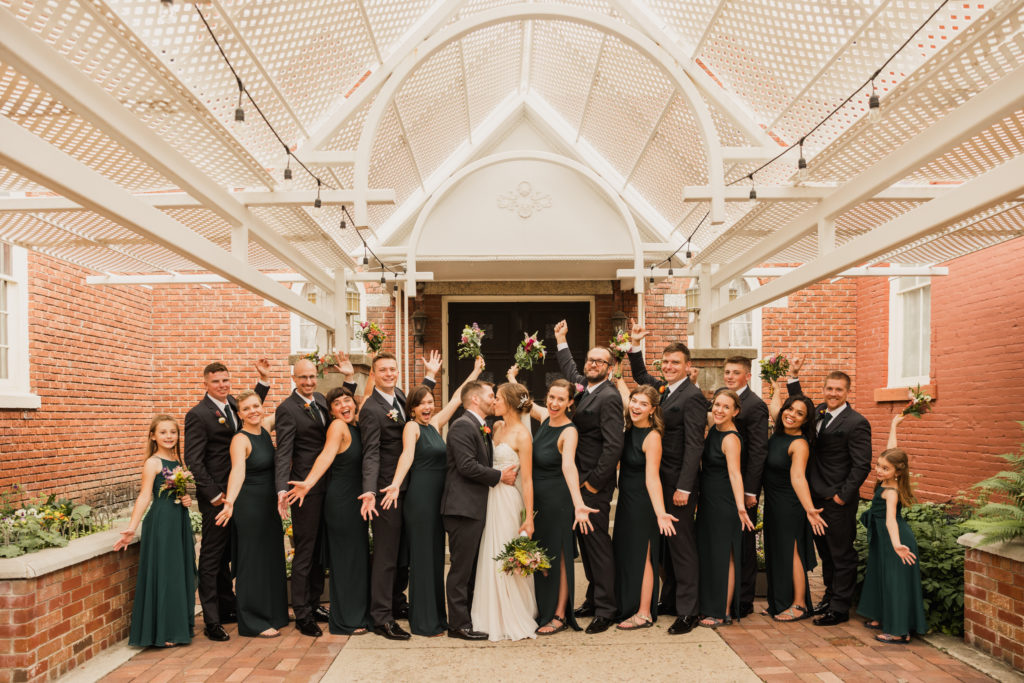 The bridal party smiles excitedly as the bride and groom kiss for this group shot, taken in front of the Hearthside Event Center by Robin Kunzler Photo