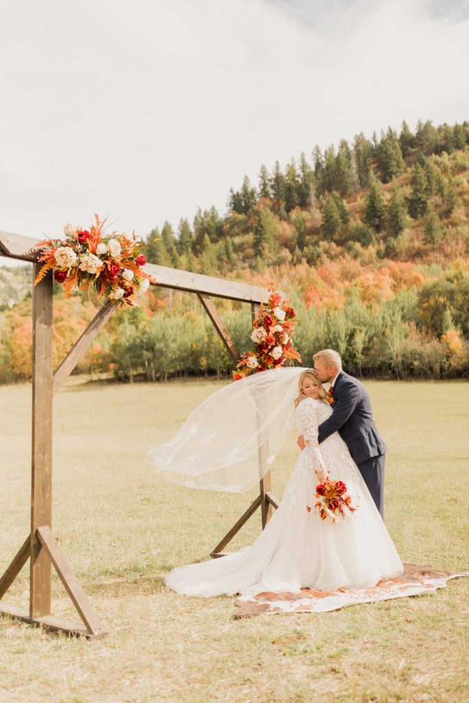 Groom planting a kiss on bride's temple as they pose next to their wedding arch, captured by Robin Kunzler Photo